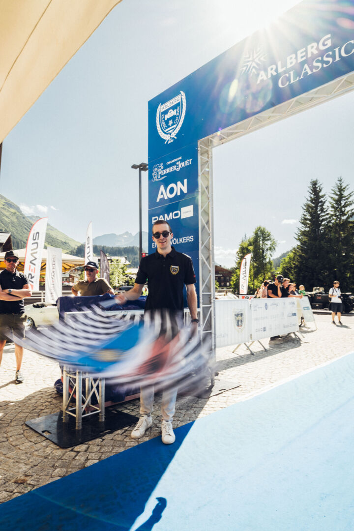 Florian Hitschfel schwenkt eine Zielflagge unter dem Start- und Zielbogen der Arlberg Classic Car Rallye in Lech.