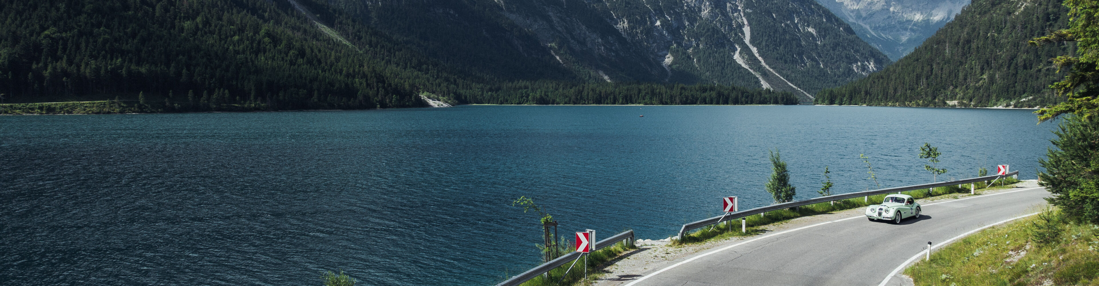 Oldtimer fährt in bergiger Landschaft mit blauen See im Hintergrund