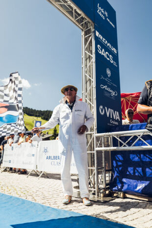 Hannes Schneider in weißem Rennanzug steht beim Startbereich des Arlberg Classic Car Rallye-Events in Lech Zürs vor einer großen Sponsorenwand und unter blauem Himmel.